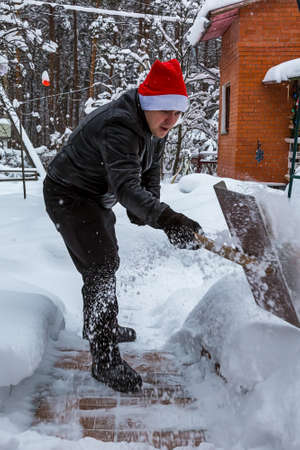 A man cleans steps from snow. In a red hat of Santa Claus and a leather black jacket. In felt boots. Clearing snow from the backyard after a snowfall.の写真素材