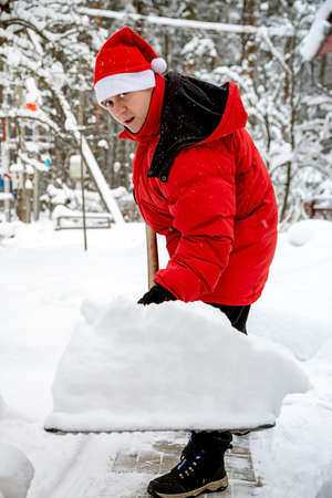 man in red down jacket and red hat of Santa claus clears snow in backyard. Clears snowdrifts on path to home. In snowfall. Man remove snow with big plastic shovel with wooden handleの写真素材
