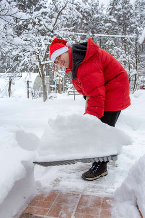 man in red down jacket and red hat of Santa claus clears snow in backyard. Clears snowdrifts on path to home. In snowfall. Man remove snow with big plastic shovel with wooden handleの写真素材