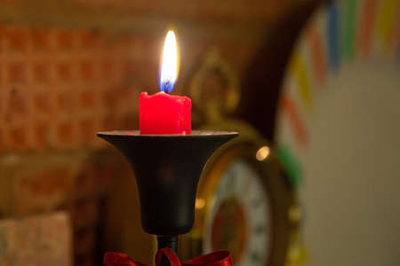 Burning red candle in black candelabra. Stands on brick fireplace. Clocks on backgroundの写真素材