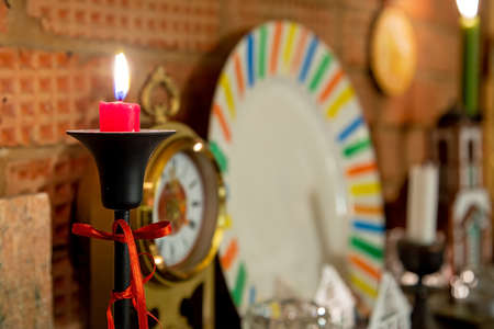 Burning red candle in black candelabrum tied with red ribbon. Stands on brick fireplace. Old retro clocks and white plate on backgroundの写真素材