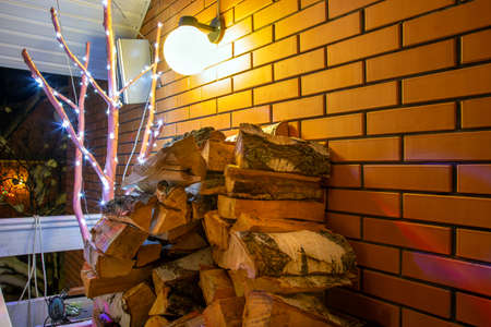 Pile of birch firewood for fireplace. Red brick wall and lantern is burning on background. Winter nightの写真素材