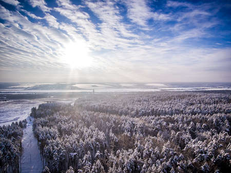 Aerial view on snow winter forest and field near road. Frozen river on background. Clouds with sun in blue skyの写真素材