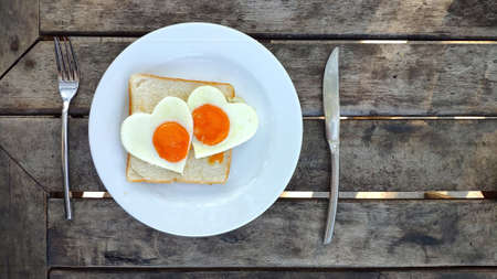 Breakfast for Valentines day. Fried eggs in the shape of a heart at toast on white plate and wooden table on the beach.  Knife and fork lie nearbyの写真素材