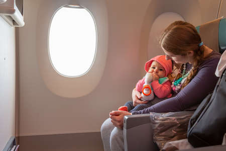 Mother carry her infant baby during flight. Sitting together near the window in airplaneの写真素材