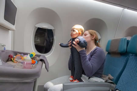 Infant plays on mother hands at the airplane.  Amazed of tablet monitor and flight.  Plane baby bassinet on the foregroundの写真素材