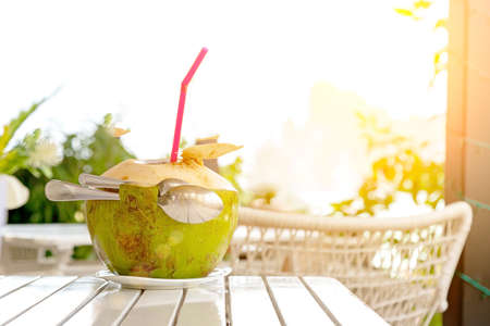 Fresh coconut juice with straw and two spoons on white wood table against blurred beach and mountain
