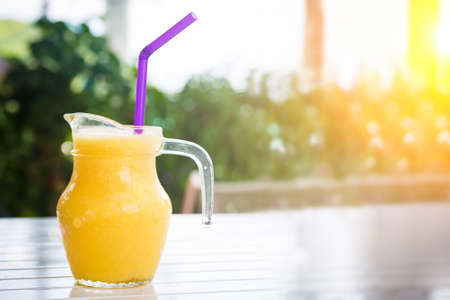 Fresh orange juice in glass in the shape of a jug with a purple straw. Smoothie on the white wooden table.の写真素材