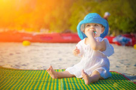 Little baby girl 8 month is sitting on the sandy beach in blue hat and white dress. She is impressed of the sea.の写真素材