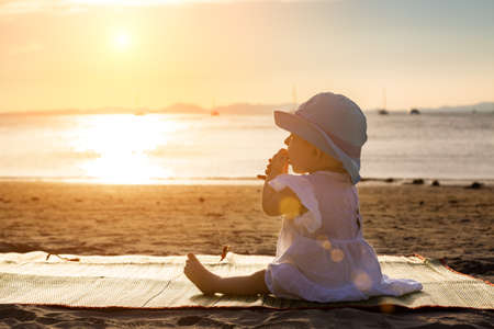 The baby is teething. Everything gnaws, chewing. Infant girl sitting on the tropical sand beach and looking for sunsetの写真素材
