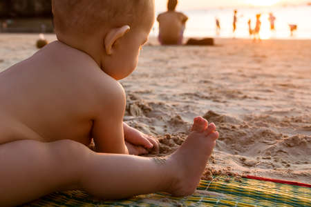 Baby playing and eat sand on the tropical beach. Infant girl of nine month.  Sunset light. Golden hour.の写真素材
