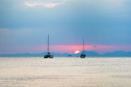 Three sailing yachts in the sea. During sunset, the sun sets over the mountains.