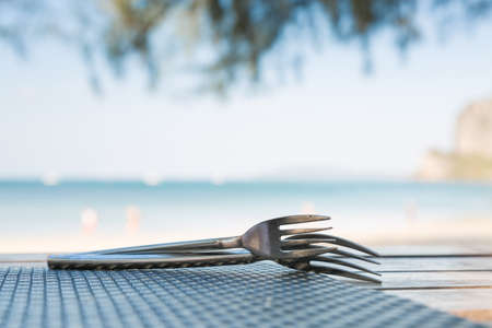 Two forks lie on top of each other. On a wooden table with bamboo mat. On the background of the sea and sandy tropical beach in blurの写真素材