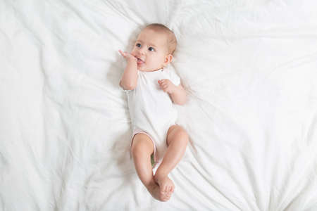 The baby teething Close up portrait of little baby girl in white lying on a white bed. puts her hands in her mouthの写真素材