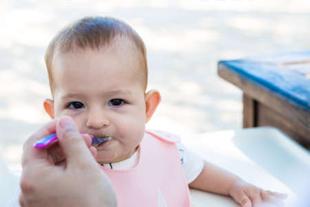 Infant girl eating porridge with a plastic spoon on the street. Baby in In a special rubber apron.の写真素材