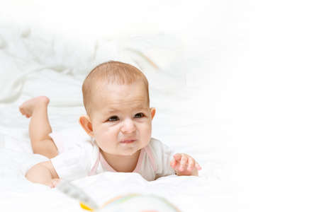 Sad and disappointed baby girl in white clothes is lying on a white bed and looking at right, Infant...