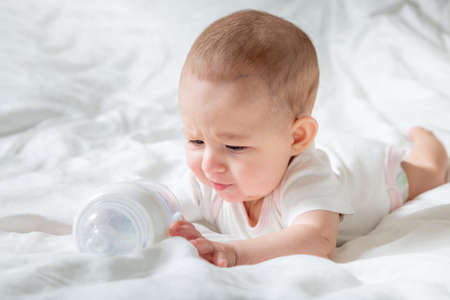Sad and crying Infant baby girl lying on the white bed with special bottle of water with nipple....