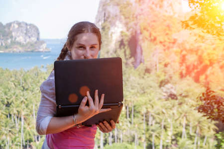 Freelancer girl hiding behind a laptop screen. She stands on the edge of a rock with its back to the sea and tropical forest landscape.の写真素材