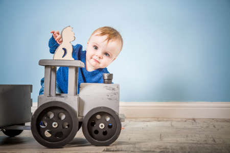 Happy boy blond in a blue sweater sits on a wooden floor. One year old baby playing with wooden toys. Cat and train made of wood.の写真素材