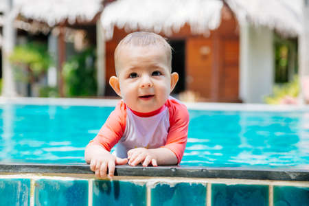 A cute and happy little baby holds to the side of the tropical pool and looks to the camera and laught. Infant girl in swim suit at summer vacationの写真素材