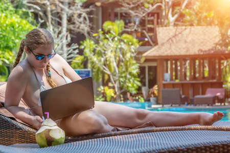 A young girl sits on a lounger by the pool. Sunbathes and works for the laptop. Sunny day.の写真素材