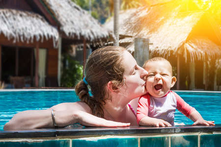 Happy young mother with a small infant baby in the pool outdoors. A woman kisses her joyful, opened his mouth and laughing child.の写真素材