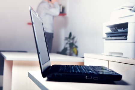 Laptop on the table in the office. Against a background in blur, a young businessman is talking on...