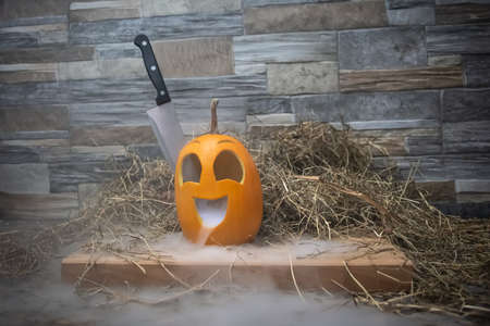 Yellow and funny halloween pumpkin with a knife in his head and smoke or steam from his mouth. Stands on a wooden stand against a stone wallの写真素材