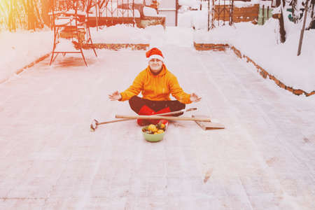 A man in a red Christmas hat and boots, in a orange jacket, sits in a lotus position on the site of his yard with a brush and a shovel. He cleaned the snow from the backyard and was pleased with it.の写真素材