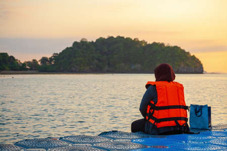 A woman in a bright orange life jacket sits on a pontoon pier early in the morning during sunrise....