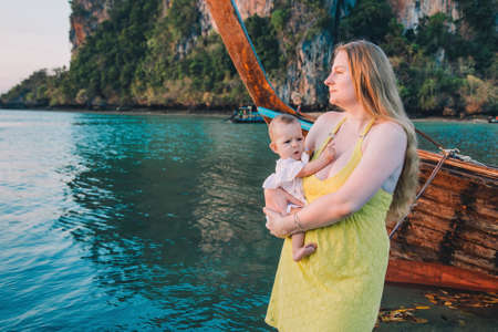 Pensive mother with a disgruntled baby stand on the pier against the background of water in the early morning and meet sunrise in anticipation of the boat.の写真素材