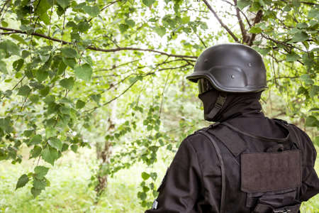 A soldier in special black equipment. Warrior without insignia and stripes. Special forces in the woods aiming with weapons. Tactical exercises.の写真素材