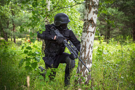 A soldier in special black equipment. Warrior without insignia and stripes. Special forces in the woods aiming with weapons. Tactical exercises.の写真素材