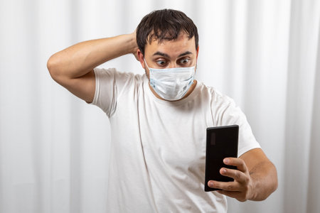 A man in panic looks at the phone and reads the news of the epidemic. In a protective medical maskの写真素材