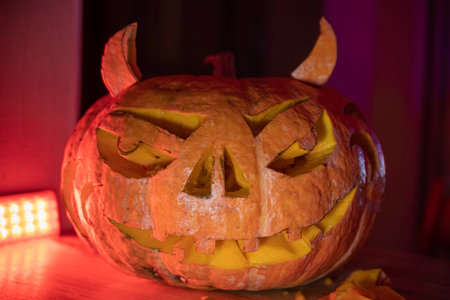 on the table there is a carved Halloween pumpkin with horns and a red backlight on the backgroundの写真素材