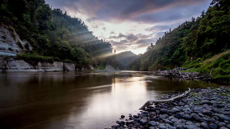 Wide river with stones on the shore, near the mountains with green trees on a cloudy eveningの写真素材