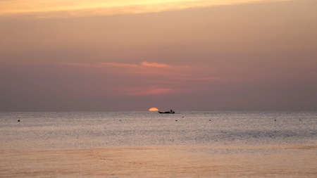 A fishing boat off the coast at sunset in Africa with part of the setting sun over the horizonの写真素材