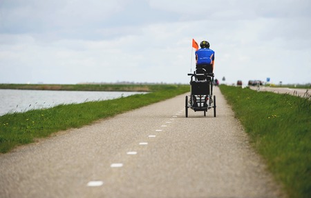 AMSTERDAM, NETHERLANDS, April 2017: Man on the Bike Pulling Baby Cart for a Bicycle. Bicycle path with Lake on One Site and the Motor Road on the Other.のeditorial素材