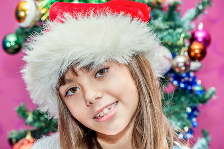 Happy child girl in a Christmas hat standing in the front of Christmas treeの写真素材