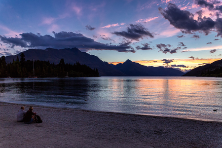 People sitting by the lake Wakatipu and watching beautiful sunset in Queenstown, New Zealandの写真素材