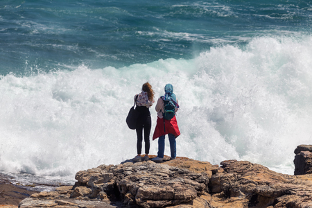 Two young female tourists facing and photographing a huge dangerous wave in Hermanus, South Africaの写真素材