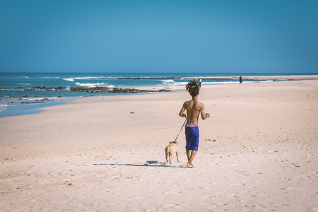 African man with dreadlocks hair running with his dog on the beach of Jeffreys Bay, South Africaの写真素材
