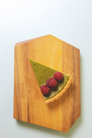 A piece of matcha cheesecake tart on a wooden plate topped with raspberriesの写真素材