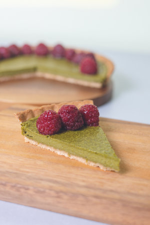 A piece of matcha cheesecake tart on a wooden plate topped with raspberriesの写真素材