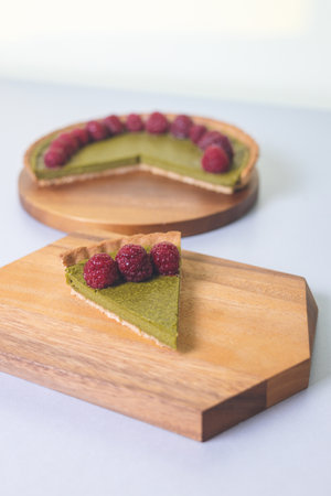 A piece of matcha cheesecake tart on a wooden plate topped with raspberriesの写真素材