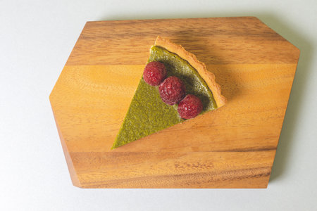 A piece of matcha cheesecake tart on a wooden plate topped with raspberriesの写真素材