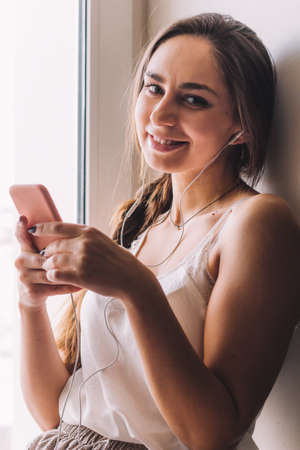 beautiful young woman sitting on the windowsill listen to music hold a smartphoneの写真素材