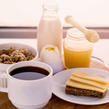 healthy breakfast fresh cereal, cheese sandwich, hard boiled egg, milk bottle, coffee cup and honey jar, toned warm sunny morning lightの写真素材