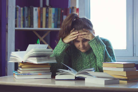 sad woman in a green sweater at a table in the middle of a stack of books and study materials, a...