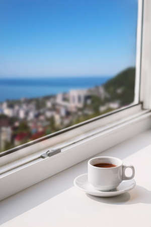 white cup with drink on a window sill with blurred city view with  blue sky and sea copy space